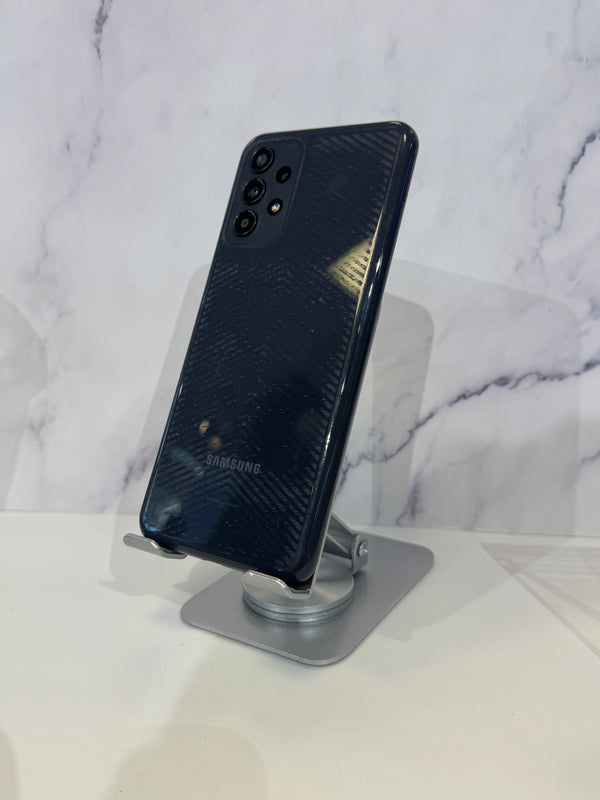 A Samsung smartphone with a dark blue textured back panel, displayed on a silver stand against a white marble-patterned background.
