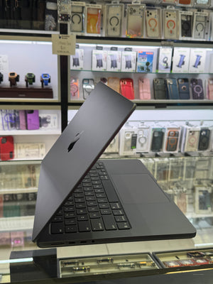 A dark gray Apple MacBook laptop with a black keyboard cover, shown open on a retail display counter with shelves of phone cases and accessories in the background.