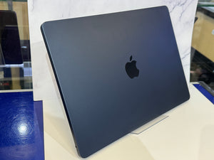 A closed Apple laptop in dark blue-gray, shown standing upright on a display stand in a showroom.