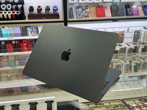 A closed dark gray Apple MacBook laptop is shown from the back, resting on a glass display counter inside a retail store with shelves of phone cases and accessories in the blurred background.