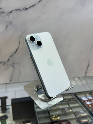 A light blue-gray Apple iPhone with a silver bumper and dual rear cameras, displayed on a metallic stand in front of a marble-patterned wall.