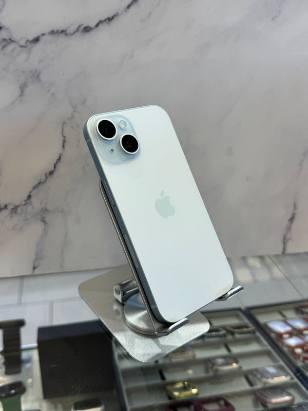 A light blue-gray Apple iPhone with a silver bumper and dual rear cameras, displayed on a metallic stand in front of a marble-patterned wall.