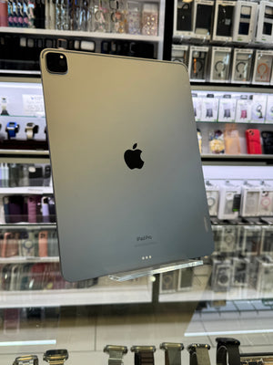 A silver Apple iPad Pro standing upright on a clear display stand inside a retail store, with shelves of accessories and electronics blurred in the background.