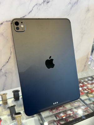 A dark gray Apple iPad Pro shown from the back, resting on a white marble-patterned surface with a blurred retail store background.