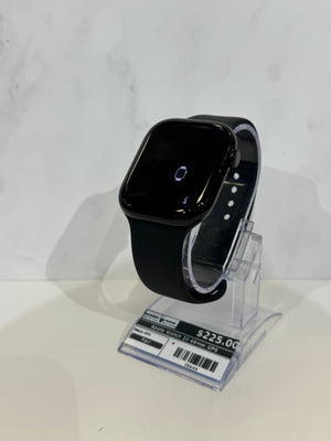 A black smartwatch with a glossy finish and black band, displayed on a clear acrylic stand with a price tag attached. The background is a clean white marble-patterned wall.