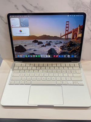 A silver aluminum laptop with a white keyboard and trackpad, shown open on a light surface against a white marble-patterned background. The screen is on and displays a macOS desktop with the Golden Gate landscape wallpaper.
