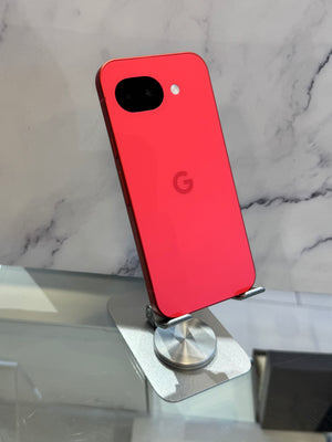 A red Google Pixel smartphone shown from the back, resting on a small silver display stand against a white marble-patterned background.