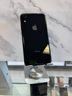 A black Apple iPhone XR standing upright on a silver metal display stand, showing the back of the phone with the Apple logo and iPhone text. The phone is photographed indoors against a white marble-patterned wall, with a blurred watch display case visible in the lower right background.