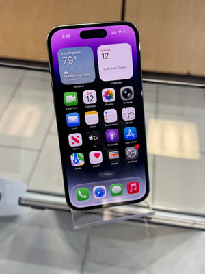 A black iPhone with a purple and pink iOS home screen, displayed on a clear acrylic stand on a reflective glass surface.