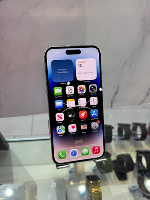 A black Apple iPhone with a large screen displaying the home screen, shown on a small display stand in a retail or showroom setting with watches blurred in the background.