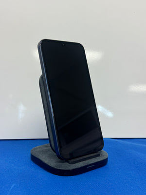 A black smartphone displayed upright on a dark gray charging stand against a white background with a blue surface below.