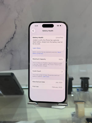 A black smartphone displayed upright on a small stand, showing the Battery Health settings screen on its home screen. The phone is photographed indoors against a light marble-patterned wall and reflective surface.