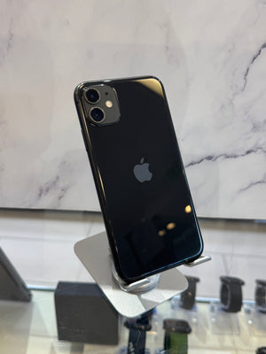 A black Apple iPhone with a glossy back is displayed on a silver stand in a retail setting, with a marble-patterned wall in the background.