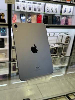 A silver Apple iPad shown from the back, standing on a glass display counter inside a retail store. The Apple logo is clearly visible in the center, with blurred store shelves and products in the background.