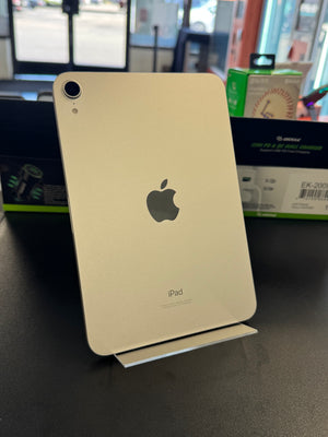 An Apple iPad standing upright on a small metal display stand on a dark tabletop, with retail packaging and a store interior blurred in the background.