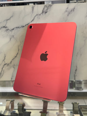 A coral pink Apple iPad shown from the back, resting on a white and gray marble surface with a store display visible in the background.