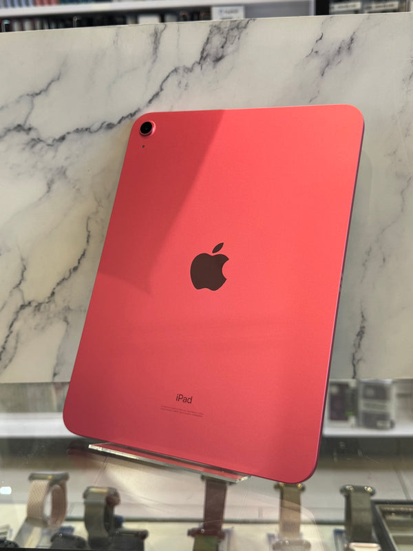 A coral pink Apple iPad shown from the back, resting on a white and gray marble surface with a store display visible in the background.
