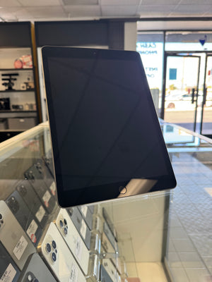 A black tablet device with a glossy screen, displayed on a glass countertop inside a retail store. Other devices are visible in the background on shelves.