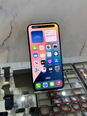A black iPhone with a colorful home screen displayed on a stand in a retail setting, with watch bands visible in the background.
