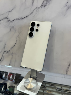 A white Samsung Galaxy Ultra smartphone shown from the back, resting on a silver metal display stand against a white marble-patterned wall.