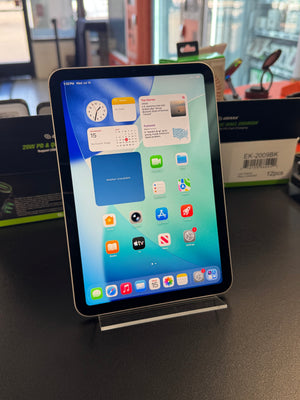 A tablet device with a black bezel and silver edges, displaying a colorful home screen, photographed on a dark surface with store fixtures in the background.