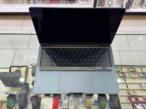 A gray Apple MacBook laptop with a black screen and keyboard, displayed on a glass counter in a watch store with various smartwatches visible in the background.