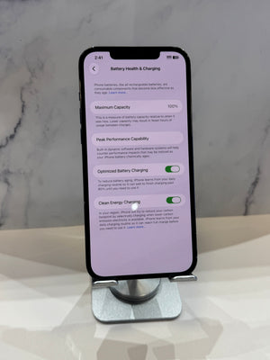 A smartphone displayed upright on a metal stand, showing the battery health and charging settings screen on a light marble surface against a marble wall.