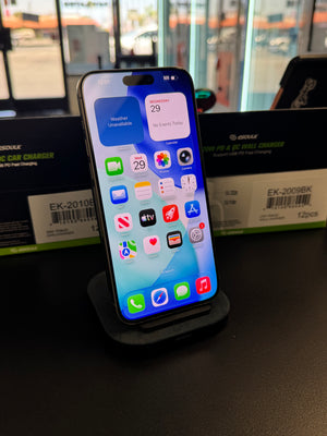 A black Apple MagSafe-compatible wireless car charger docked on a smartphone stand, with its retail packaging boxes visible in the background on a dark tabletop.