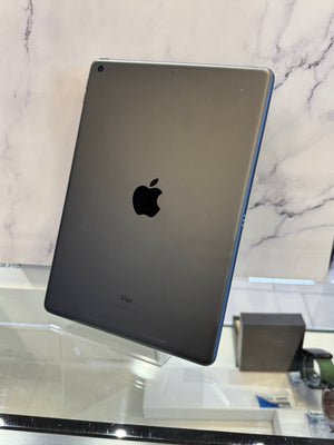 A dark gray Apple iPad Pro shown from the back, standing on a clear display stand on a glass counter with a marble-patterned background.