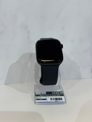 A black Apple Watch with a dark band displayed on a clear stand, with a price tag attached at the base, set against a light marble-patterned background.