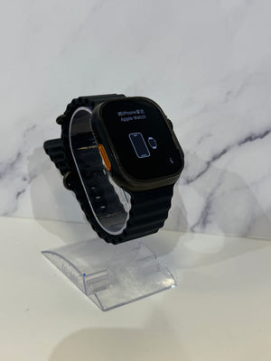 A black smartwatch with a black strap and a black rectangular display, shown resting on a clear plastic display stand against a white marble-patterned background.