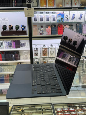 A dark gray Microsoft Surface Laptop with a matching keyboard cover is shown open in a retail store setting, with shelves of other tech accessories and watches in the background.