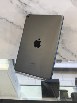An Apple iPad with a dark gray metallic back and a blue edge bumper, displayed on a clear stand in front of a white marble-patterned background.
