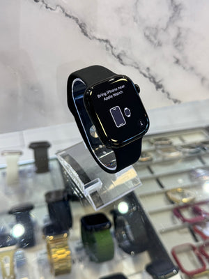 A black Apple Watch with a black sport band, resting on a clear acrylic display stand inside a watch store. The watch screen displays the 'Bring iPhone next' interface, and the background shows a marble wall and other watch displays.