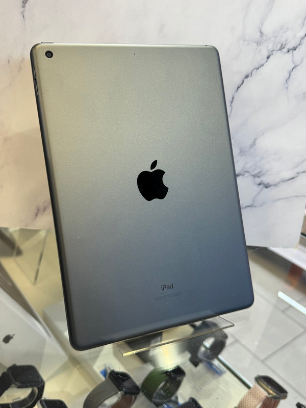 A silver Apple iPad shown from the back, resting on a clear display stand in front of a white marble-patterned background.