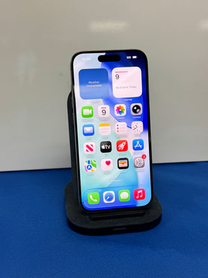 A black iPhone with its screen on, resting on a black charging dock against a white wall and blue surface.
