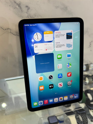 A black tablet, likely an iPad, displaying its home screen with various app icons and widgets. The tablet is resting on a glass display counter in front of a light marble-patterned wall.