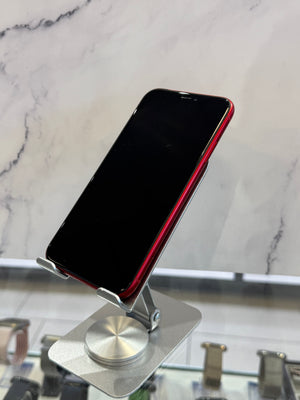 A black smartphone with a red side bumper is mounted on a silver metal display stand against a white marble-patterned background.