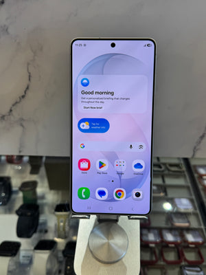 A Samsung Galaxy smartphone displayed upright on a metal stand in a retail setting, with a marble-patterned background and watch displays below.