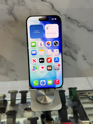 A black iPhone with its screen on, resting on a silver metal display stand. The background is a white marble-patterned wall with other watch displays visible in the blurred foreground and background.