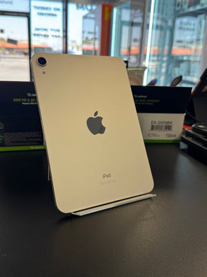 A gold Apple iPad standing upright on a small display stand on a dark tabletop, with the back of the device visible and the Apple logo centered. The background shows a blurred retail or office interior with windows and boxes.