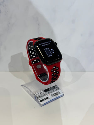 A smartwatch with a red and black perforated sport band displayed on a clear stand against a light marble-patterned background.