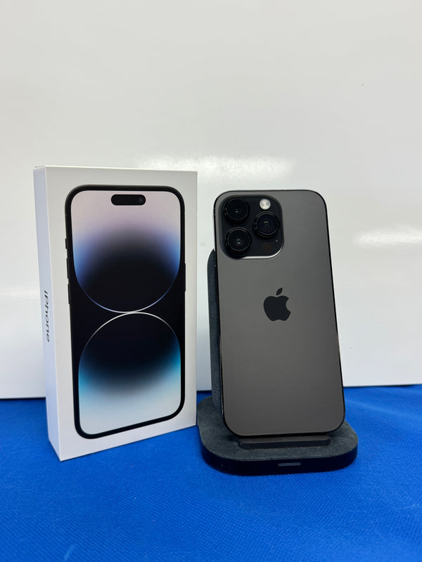 A dark gray Apple iPhone 15 Pro shown from the back on a black charging stand, next to its white retail box, set against a white background and blue surface.