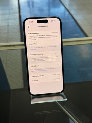 A smartphone displayed upright on a clear stand, showing the Battery Health screen on the home screen. The phone is photographed in a real indoor setting with a reflective dark surface and blue-toned background.