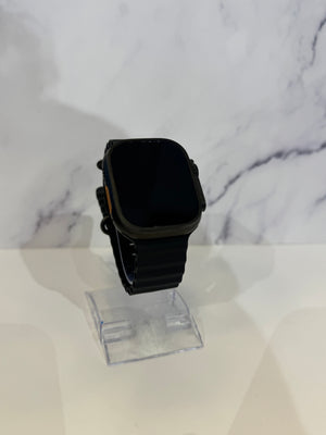 A black smartwatch with a rectangular face and black band, displayed on a clear acrylic stand against a white marble-patterned background.
