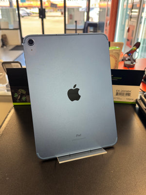 An iPad Pro standing upright on a small display stand on a dark tabletop, with the Apple logo visible on the back. The background shows a store or office interior with boxes and equipment.