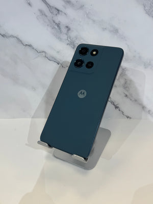 A dark teal-blue Motorola smartphone shown from the back, resting on a small clear display stand against a white marble-patterned background.
