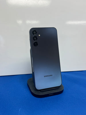 Samsung smartphone shown from the back, resting on a black charging stand against a white wall with a blue surface below.