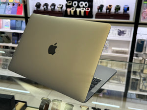 A closed Apple MacBook laptop in metallic gold is shown at an angle on a glass display counter, with a blurred watch store background.