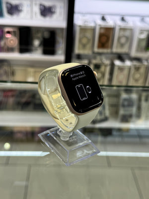 A smartwatch with a cream-colored body and a black rectangular display screen, mounted on a clear plastic display stand in front of a blurred glass display case.